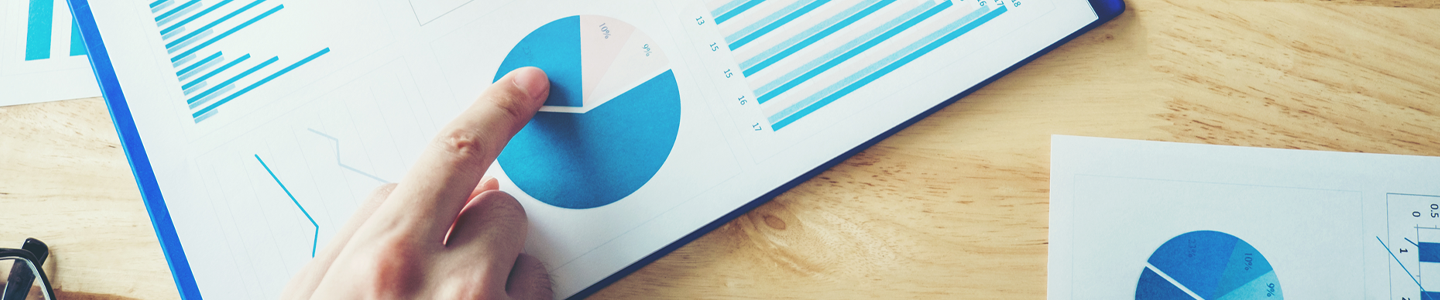 A hand pointing to a pie chart on a report resting on a table with other reports and a pair of glasses.