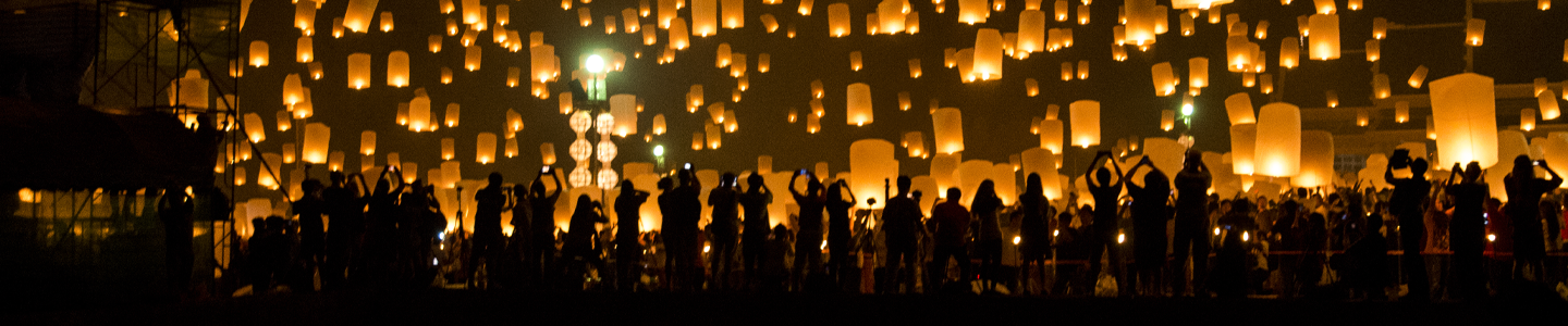 Silhouette of a crowd of people taking photos of amber-colored floating lanterns above their heads with the reflection in water behind them.