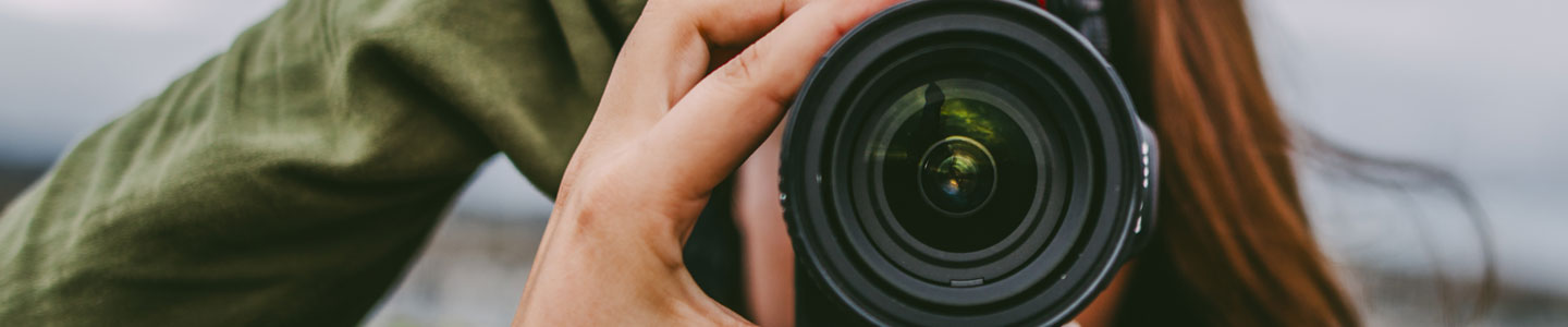 Woman in a green jacket holding a camera that's covering her face at a waterfront.