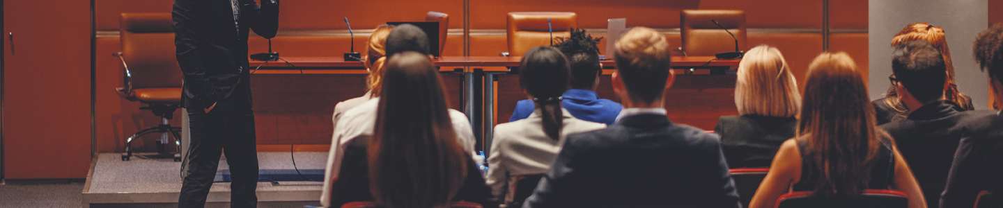 A man in a suit with his hand in his pocket, holding a microphone and speaking to people in chairs in a conference room.