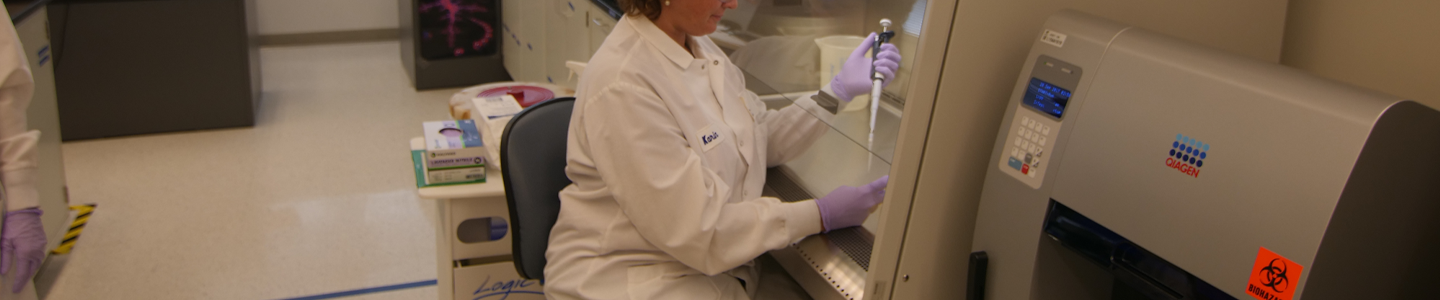 Side view of female ATCC scientist wearing lab coat, safety glasses, and gloves, holding a pipette with plunger under a safety hood in a lab.