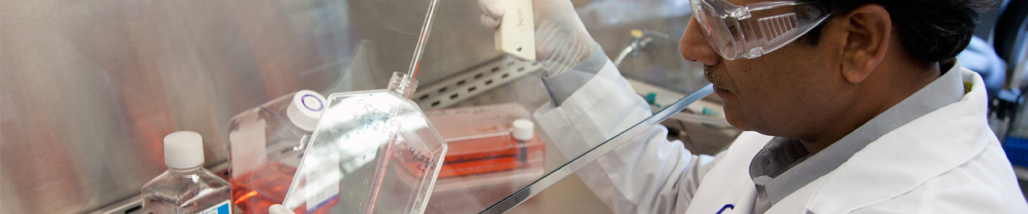 ATCC scientist in lab coat, safety glasses, and gloves, holding pipette controller and flask under hood in lab.