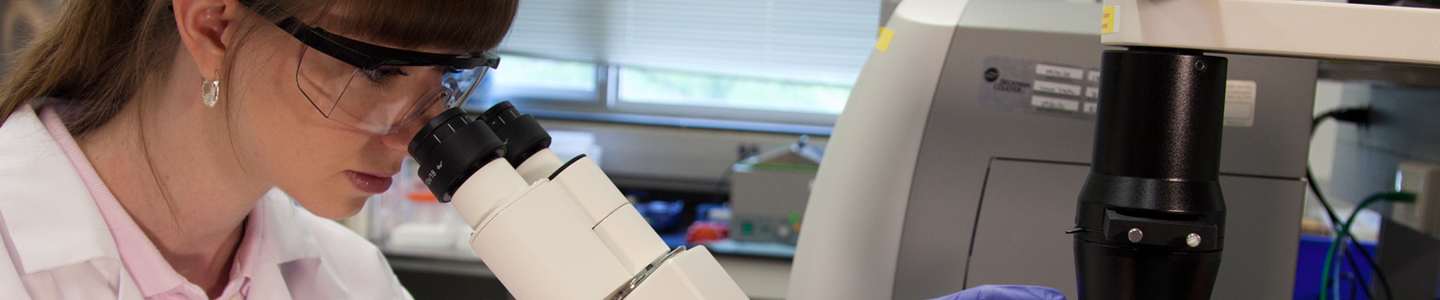 Female ATCC scientist in lab coat, safety glasses, and gloves, looking at media in a flask using an inverted microscope.