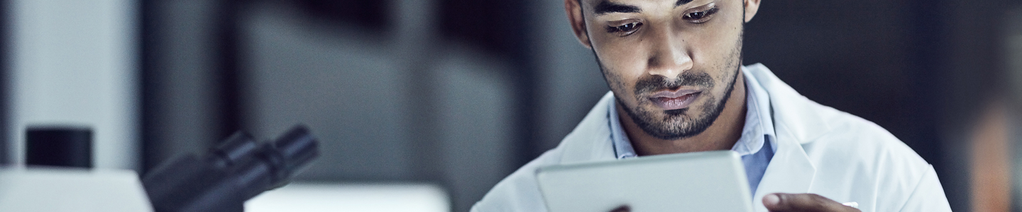 Male scientist in white lab coat, sitting next to microscope, looking at tablet.