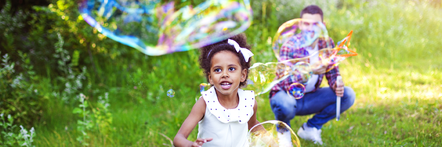 A young Black girl with a ribbon in her hair running outside in the grass toward a huge elongated bubble while a smiling man with a bubble blower squats in the background.