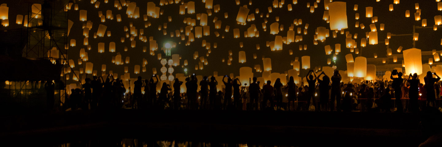 Silhouette of a crowd of people taking photos of amber-colored floating lanterns above their heads with the reflection in water behind them.