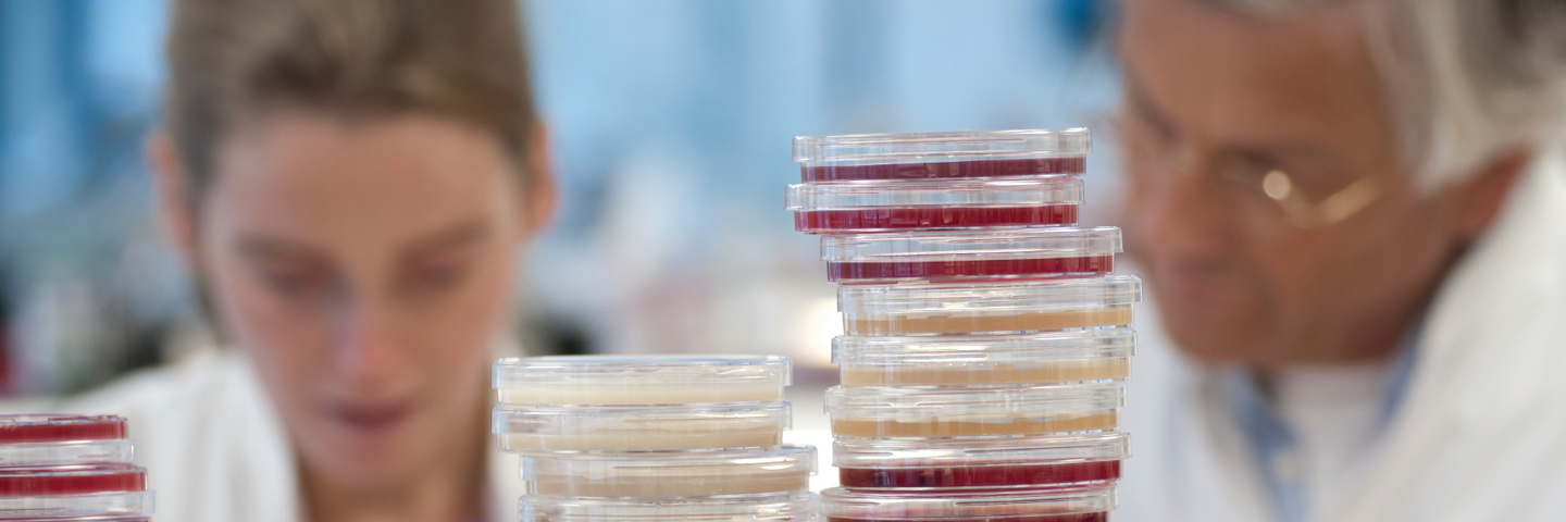 Scientists in lab coats seen over top of stacks of agar plates and small vials.