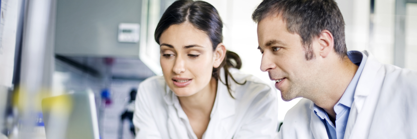 Female and male scientist in lab coats looking at laptop in a lab.