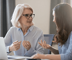 A gray-ha+F422:F464ired woman in glasses talking to a dark-haired woman at a desk in front of a laptop.