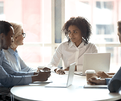 Young, black female in white shirt talking to others around a table in a conference room.