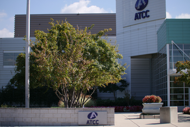Exterior of ATCC office building with ATCC logo and "ATCC" on building above entrance, and tree and shrubbery near sidewalk.