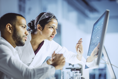 A man and woman in lab coats looking at a computer monitor with data on it.