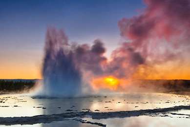 A plume of smoke rising from layers of slate-like liquid at Great Fountain Geyser in Yellowstone National Park at sunset.