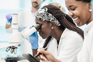 Female scientist wearing lab coat and gloves looking into a microscope.