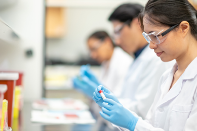 Profile of female scientist wearing safety goggles, lab coat, and gloves looking at sample in test tube, two scientist in background.