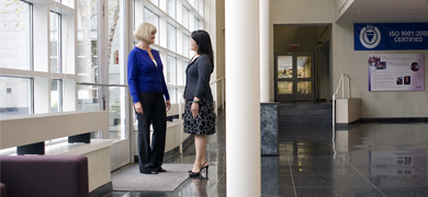 Two women in business attire standing inside office foyer near glass doors and windows.