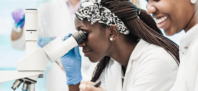 Female scientist wearing lab coat and gloves looking into a microscope.