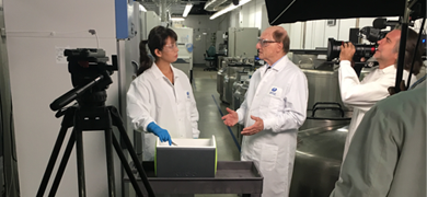 ATCC scientist with gloved hand in cooler on cart talking with Dr. Cypess, camera crew around them during filming in biorepository.