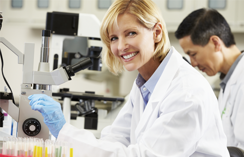 Female scientist wearing lab coat and gloves smiling at camera, at microscope holding a slide.