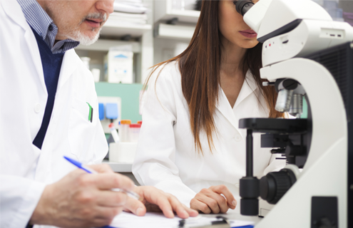 Scientist holding pen standing next to scientist looking into microscope.