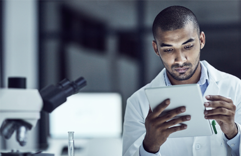 Male scientist in white lab coat, sitting next to microscope, looking at tablet.