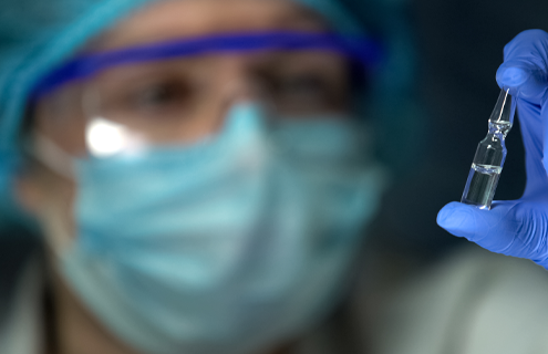 Scientist wearing cap, safety glasses, mask, and gloves holding and looking at ampoule.