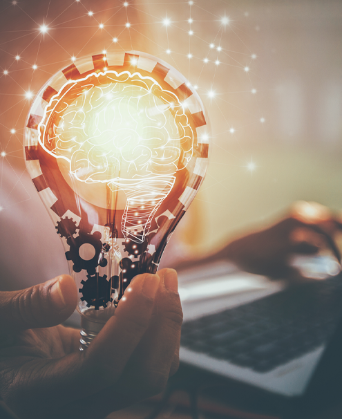 Idea-light-bulb-laptop A person sitting in the background with one hand on a computer keyboard and the other hand holding an illustration of a light bulb, with gears, filament and white outline of a human brain with sparks of light connected by lines around it.