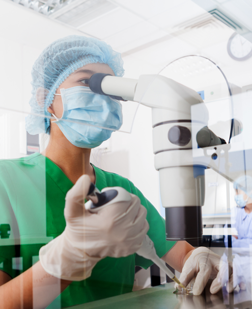 scientist-at-microscope Scientist wearing surgical smock, cap, mask, and gloves, looking into microscope while using pipette.