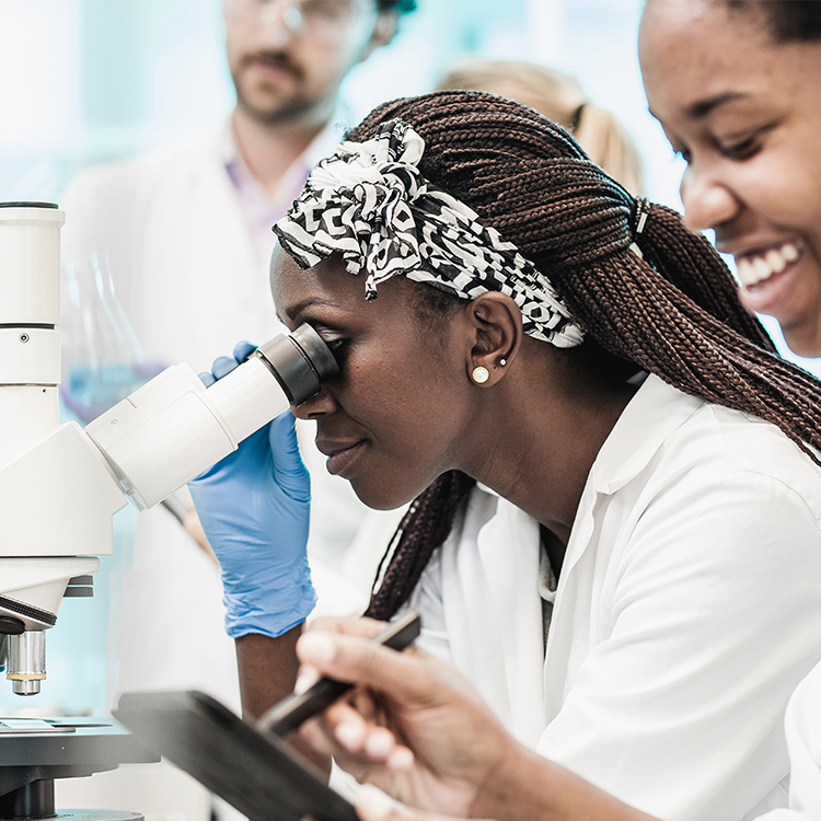 scientists-at-microscope Female scientist wearing lab coat and gloves looking into a microscope.